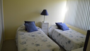A bedroom with two beds, each with a floral duvet, a bedside table with a lamp, and a window with blinds. - Cove II 233F 2 Bedroom Condo By Midnight Cove II (Siesta Key)