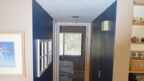 A narrow hallway with a door leading to a bedroom. The hallway has a tiled floor and a ceiling vent. There is a wicker chair in the foreground.
