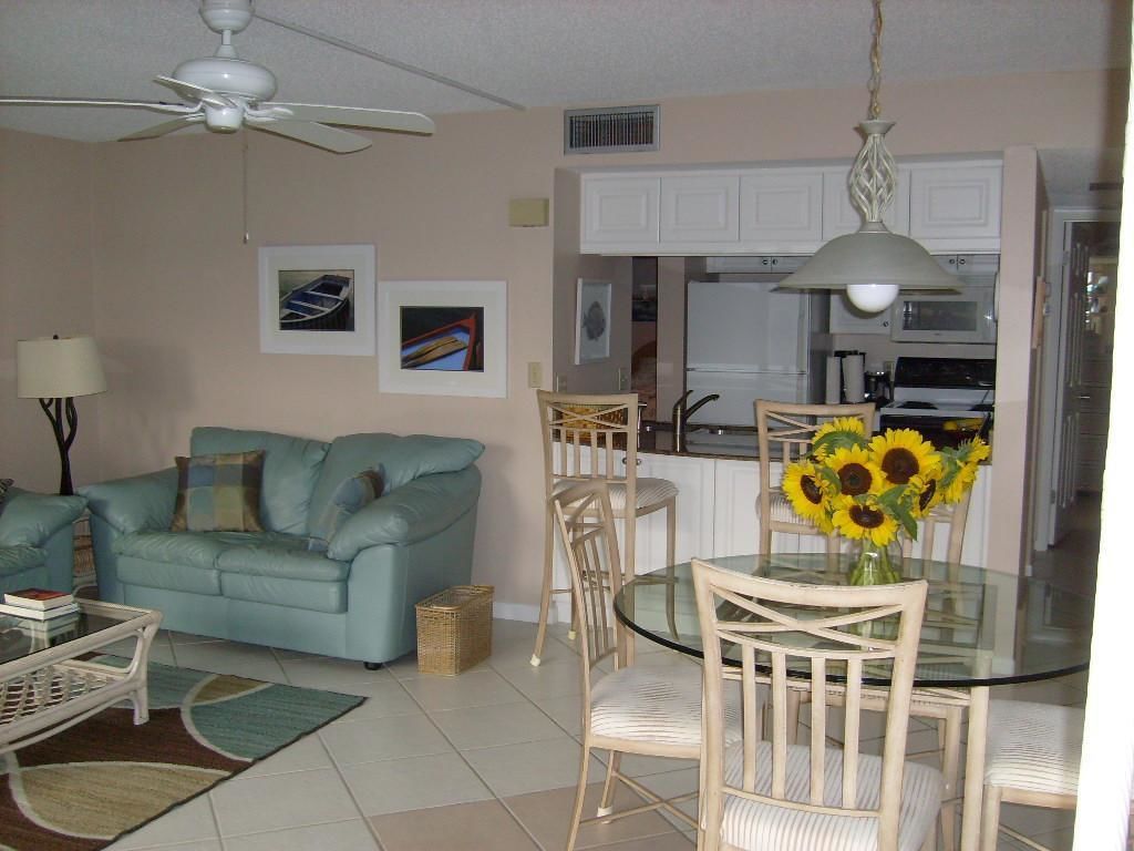 A living room with a sofa, a glass dining table with chairs, a ceiling fan, and a kitchen area with cabinets and appliances.