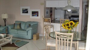 A living room with a sofa, a glass dining table with chairs, a ceiling fan, and a kitchen area with cabinets and appliances.