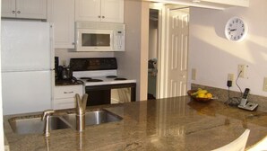 A modern kitchen with white cabinets, a granite countertop, stainless steel sink, a microwave, stove, and a clock on the wall.