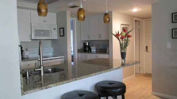 A modern kitchen with white cabinets, a granite countertop, and a stainless steel sink. There are three pendant lights hanging above the island. - Cove II 312F 2 Bedroom Condo By Midnight Cove II (Siesta Key)