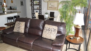 A brown leather sofa with patterned cushions, a wooden side table with a lamp, and a large green plant. - Cove II 312F 2 Bedroom Condo By Midnight Cove II (Siesta Key)