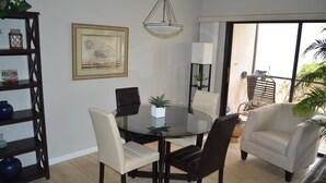 A dining area with a round glass table, two black chairs, a beige sofa, a framed picture on the wall, and a hanging light fixture. - Cove II 312F 2 Bedroom Condo By Midnight Cove II (Siesta Key)