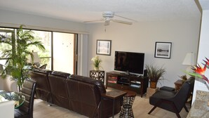 A living room with a brown leather sofa, a glass coffee table, a TV, and a ceiling fan. - Cove II 312F 2 Bedroom Condo By Midnight Cove II (Siesta Key)