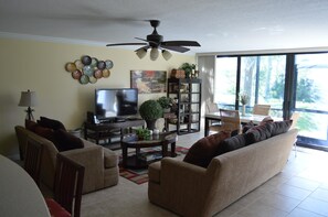 Ein Wohnzimmer mit Sofa, Sesseln, Fernseher, Deckenventilator und Bücherregal.