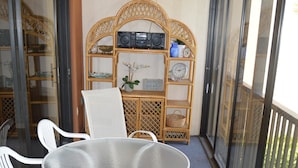 A balcony with a round table and white chairs, a wicker shelf with decorative items, and a view through the glass doors.