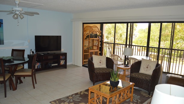 A living room with a television, wicker furniture, a coffee table, and a large window with a view of greenery.