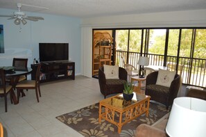 A living room with a television, wicker furniture, a coffee table, and a large window with a view of greenery. - Cove II 323F 2 Bedroom Condo By Midnight Cove II (Siesta Key)