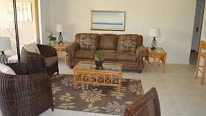 A living room with a brown leather sofa, two wicker chairs, a glass coffee table, and a floral rug.
