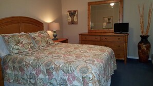 A bedroom with a bed, a dresser, a mirror, a lamp, and a vase with branches. - Cove II 510F 2 Bedroom Condo By Midnight Cove II (Siesta Key)