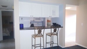 A compact kitchen with white cabinets, a microwave, a stove, and two bar stools. - Cove II 510F 2 Bedroom Condo By Midnight Cove II (Siesta Key)