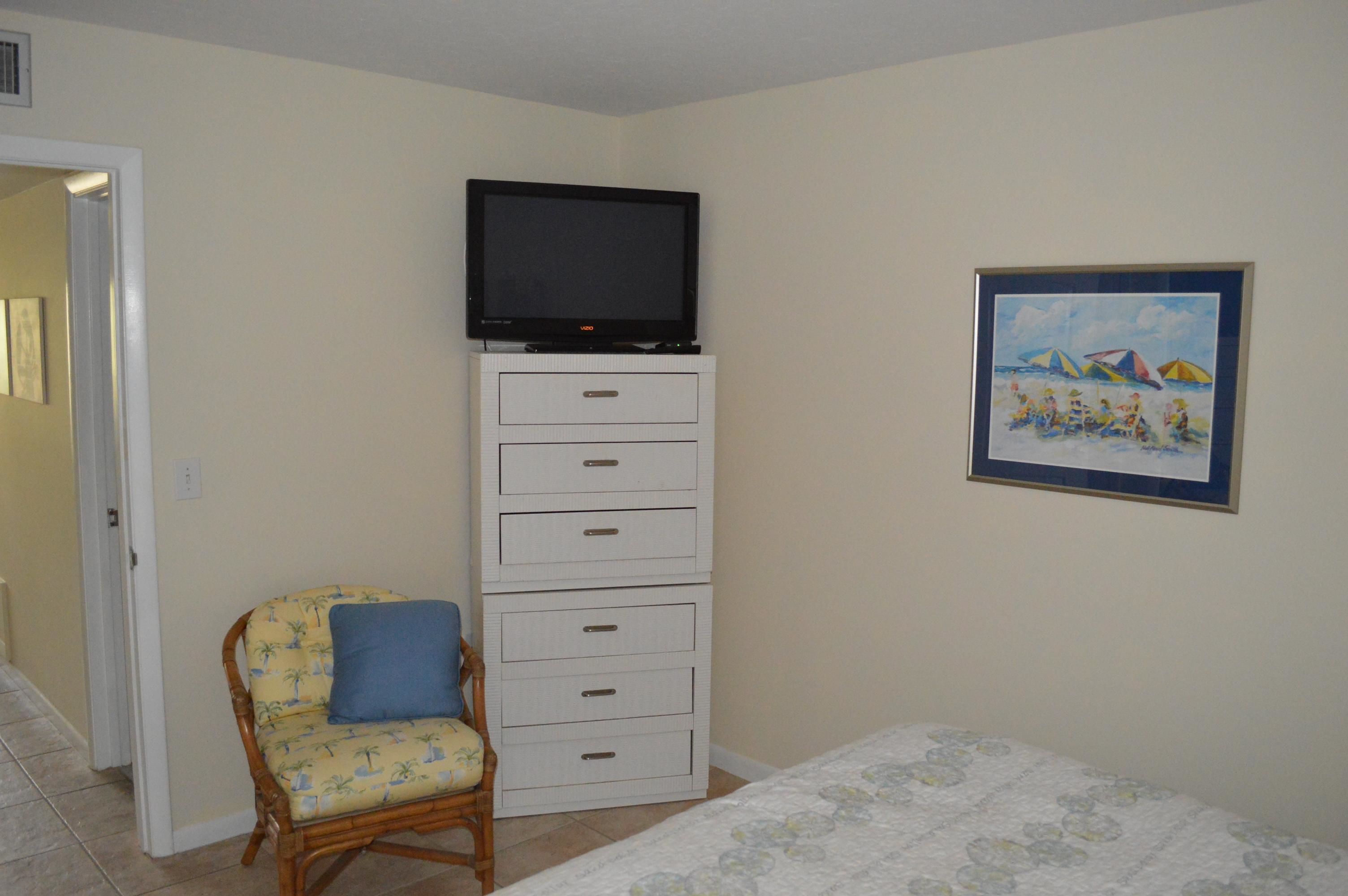 A bedroom with a bed, a television on a white cabinet, a chair with a blue cushion, and a framed beach scene on the wall.