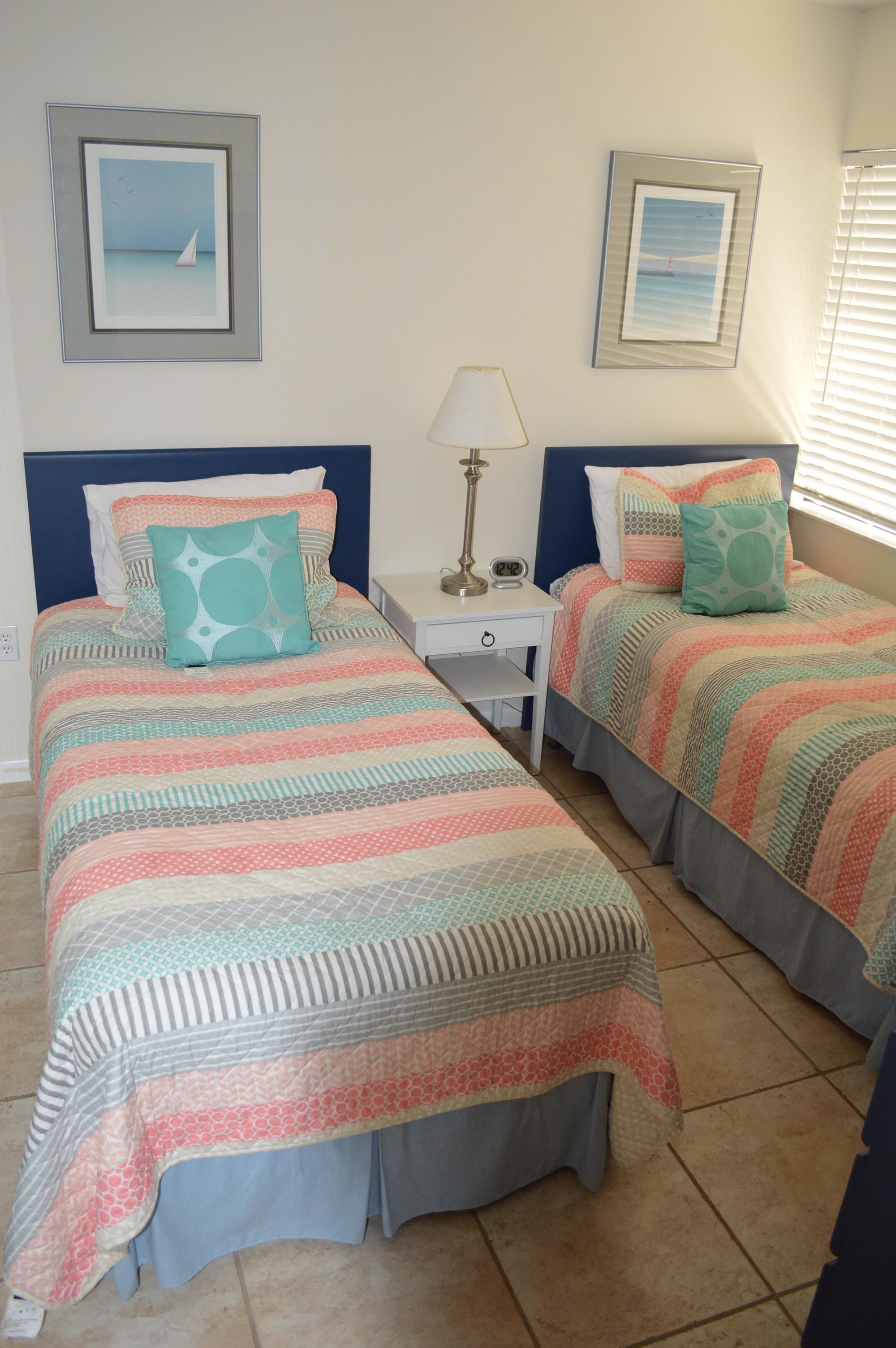 A room with two beds, each with a patterned bedspread and matching pillows. There is a nightstand with a lamp between the beds, and two framed pictures on the wall.