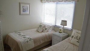 Une chambre avec deux lits, une table de chevet avec une lampe, une petite commode et une fenêtre avec des stores.