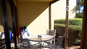 A small patio area with a round table and four chairs, a glass door leading outside, and a view of a well-maintained hedge. - Cove II 612F 2 Bedroom Condo By Midnight Cove II (Siesta Key)