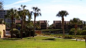 A grassy courtyard with palm trees and a building with multiple floors in the background. - Cove II 612F 2 Bedroom Condo By Midnight Cove II (Siesta Key)