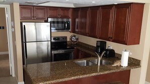 Eine kompakte Küche mit Edelstahl-Kühlschrank, Mikrowelle und Backofen. Sie verfügt über eine Arbeitsplatte aus Granit, eine Spüle und zwei Barhocker.