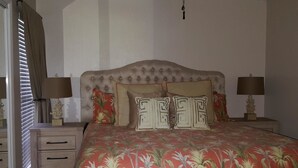 Ein Schlafzimmer mit einem gesteppten Kopfteil, einem blumigen Bettbezug, zwei Nachttischen mit Lampen und einem Fenster mit Jalousien.