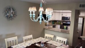 A dining area with a chandelier, a table set with a placemat, and a kitchen in the background. - Cove II 624D 3 Bedroom Holiday home By Midnight Cove II (Siesta Key)