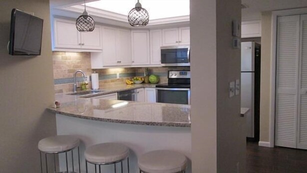 A kitchen with white cabinets, a granite countertop, a microwave, and a refrigerator. There are three stools in front of the counter. - Cove II 624D 3 Bedroom Holiday home By Midnight Cove II (Siesta Key)