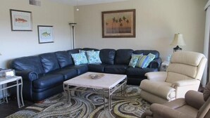 A living room with a sectional sofa, a small table, and a beige armchair. There are framed pictures on the wall and a lamp on a side table. - Cove II 624D 3 Bedroom Holiday home By Midnight Cove II (Siesta Key)