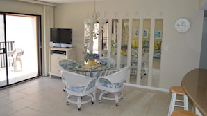 A dining area with a glass-top table, white chairs with blue cushions, a chandelier, and a television on a stand. - Cove II 633F 2 Bedroom Condo By Midnight Cove II (Siesta Key)