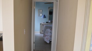 A narrow hallway with a bed in the background, a lamp on a bedside table, and a chair with a patterned cushion. - Cove II 633F 2 Bedroom Condo By Midnight Cove II (Siesta Key)