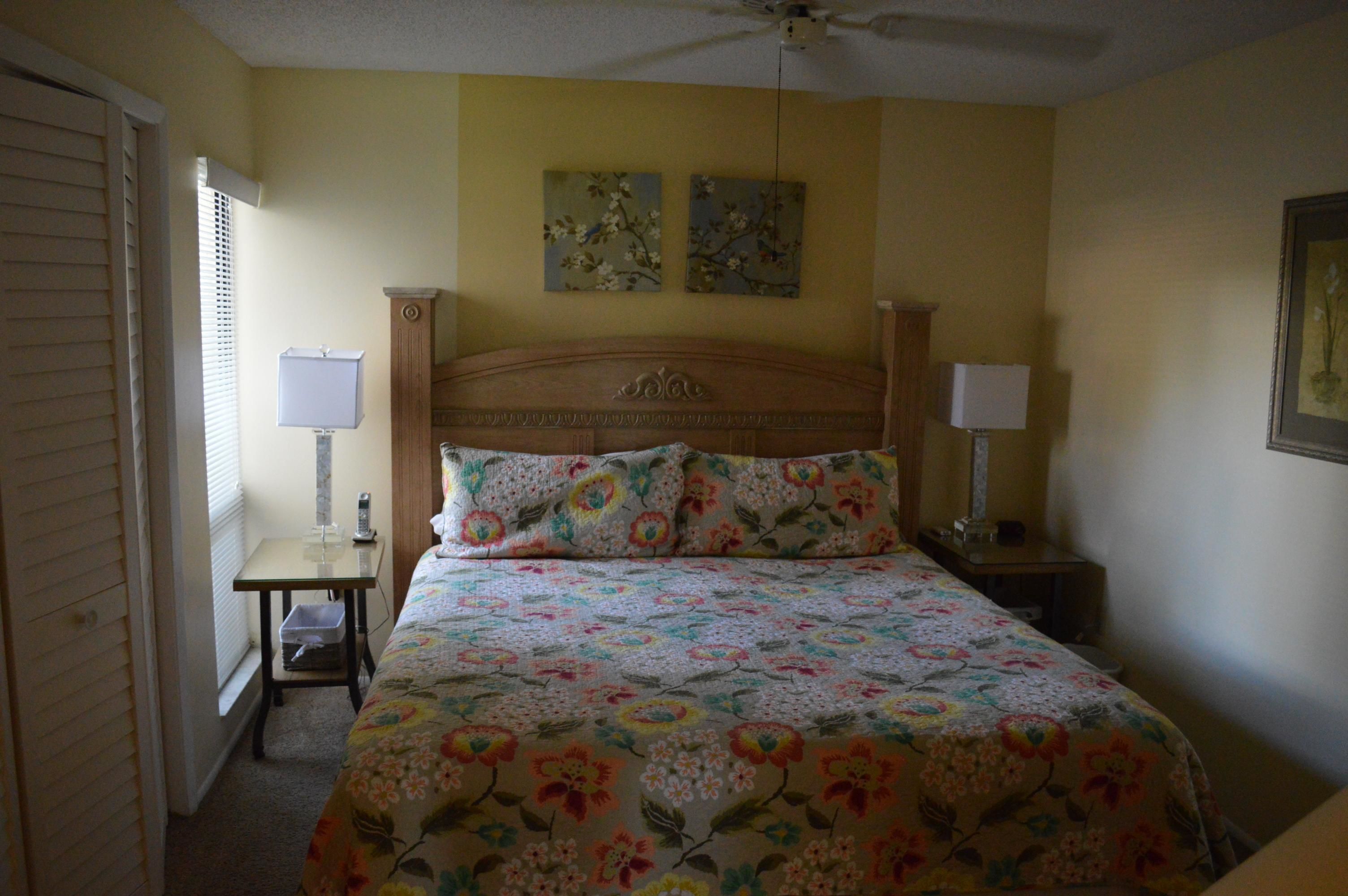 A bedroom with a floral bedspread, two bedside lamps, a ceiling fan, and two wall-mounted paintings.