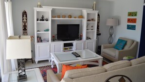 A living room with a TV, white shelves, a sofa, and a lamp. - Cove II 732F 2 Bedroom Condo By Midnight Cove II (Siesta Key)
