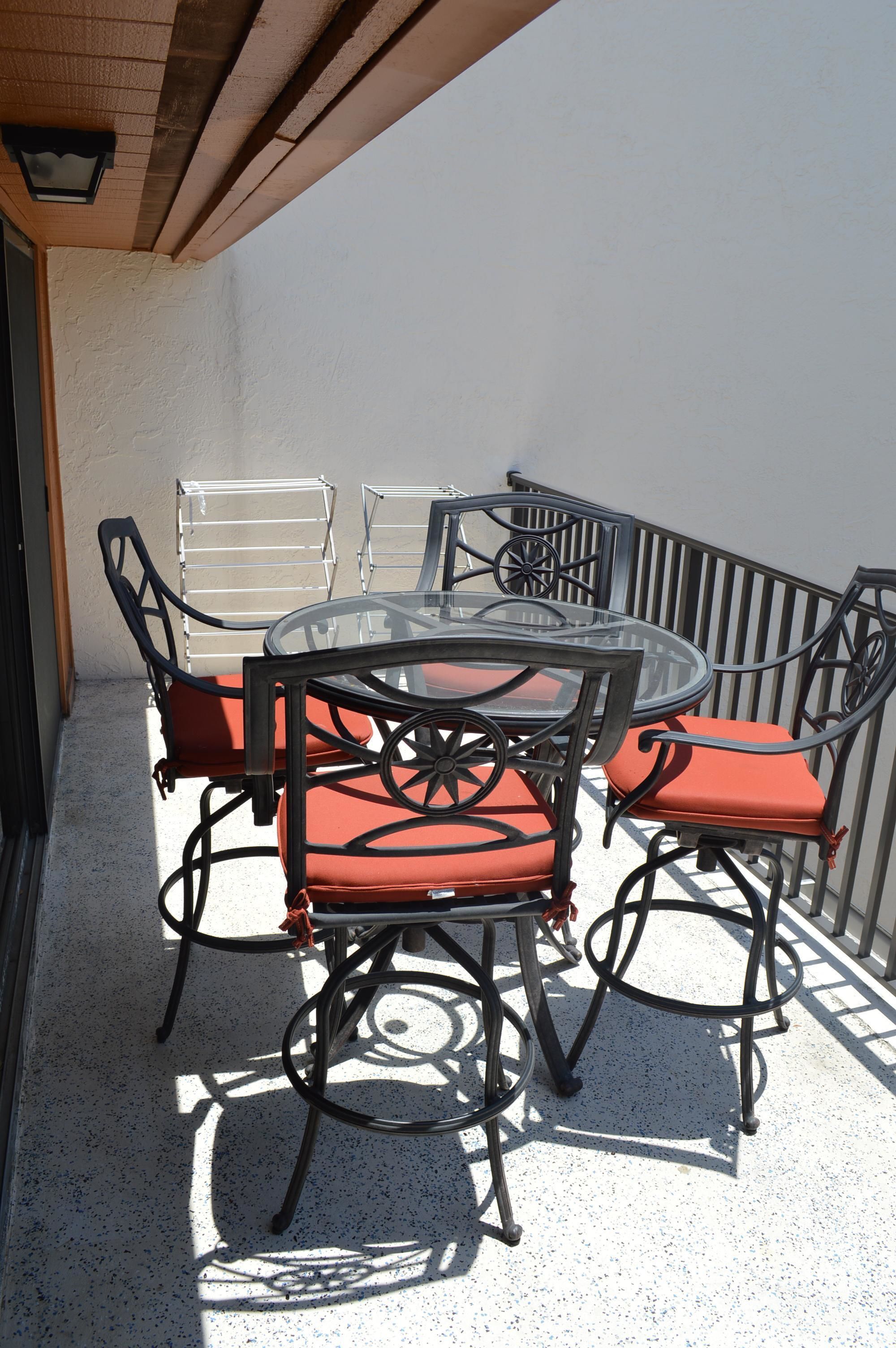 A glass-top table with four chairs, each with a red cushion, on a balcony with a metal railing and a white wall.