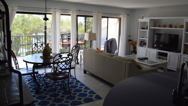 A living room with a sofa, dining table, chairs, a TV, and a balcony with a view of trees. - Cove II 732F 2 Bedroom Condo By Midnight Cove II (Siesta Key)