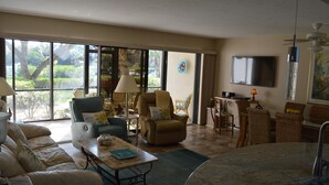 A spacious living room with a large window, a sofa, a coffee table, and a dining area with wicker chairs. - Cove II 811F 2 Bedroom Condo By Midnight Cove II (Siesta Key)