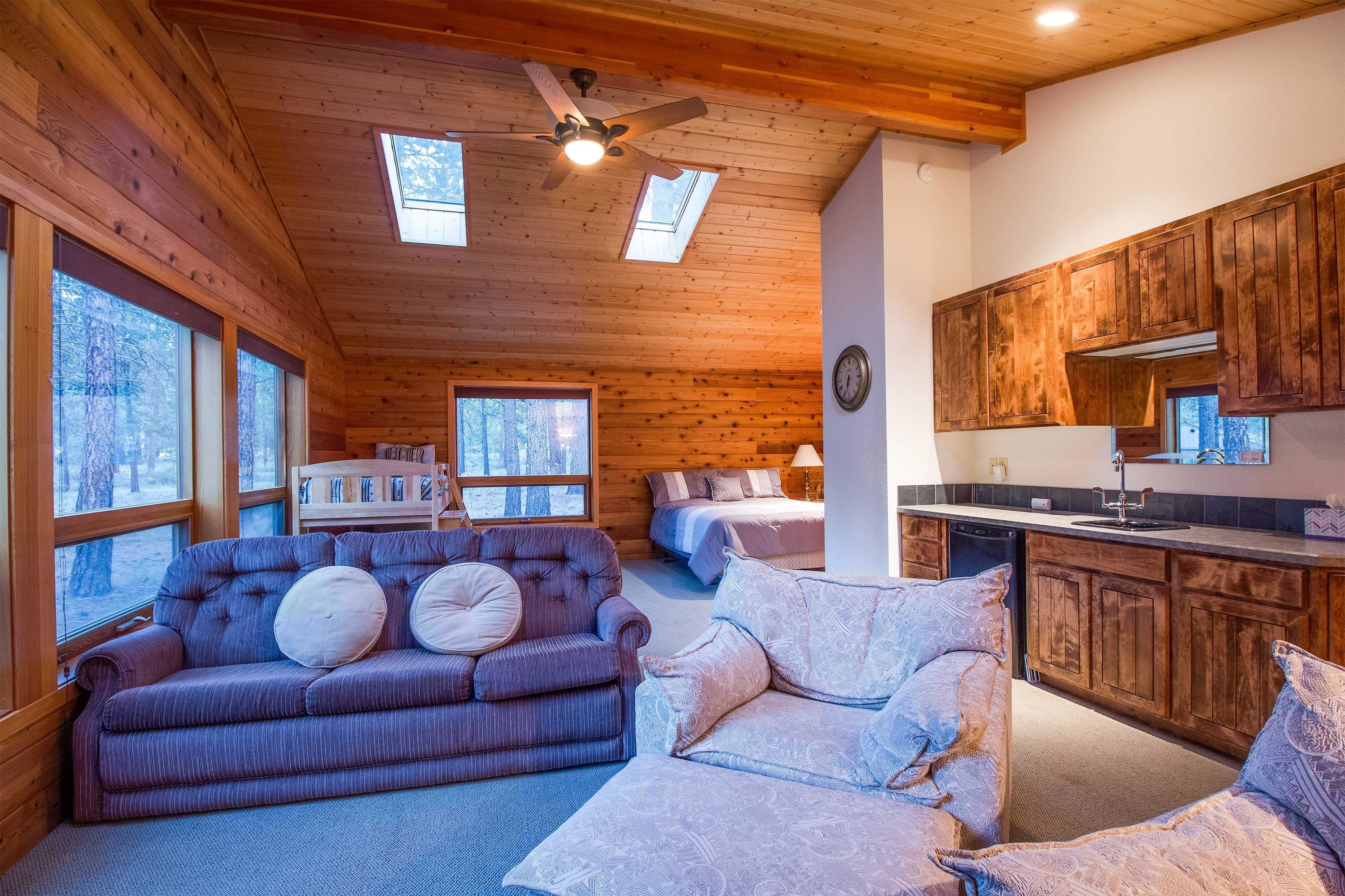 A cozy cabin interior with a wooden ceiling, a sofa, a bed, and a kitchen area.