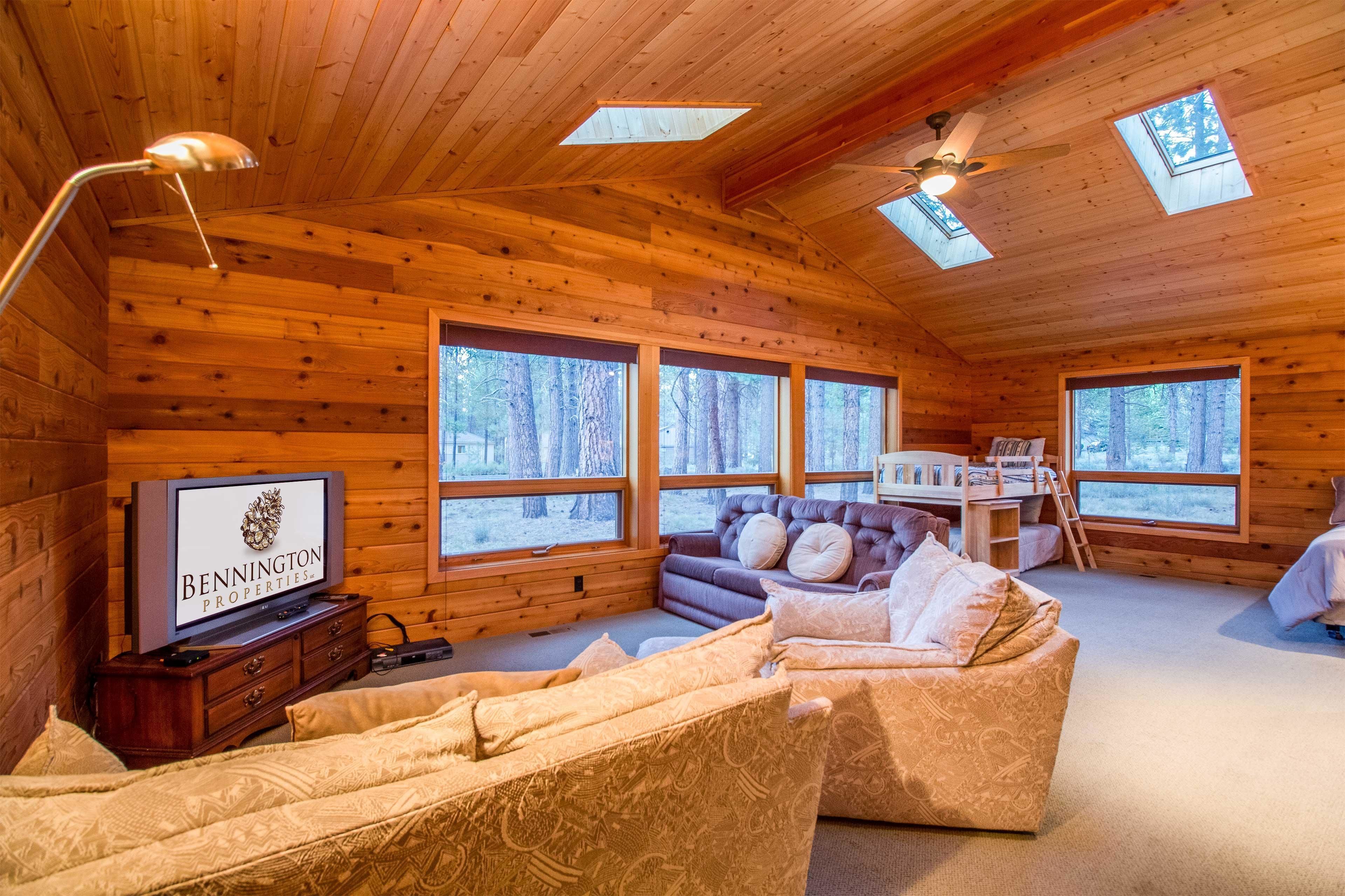 Interior acogedor de una cabaña con paredes de madera, un sofá, un televisor y ventanas amplias que ofrecen vistas al bosque.