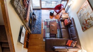 A modern living room with a leather sofa, a red chair, and a coffee table. There is a stone wall with a fireplace and a large window overlooking a deck.