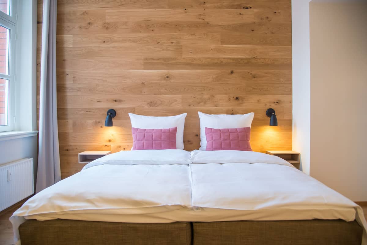 A double bed with white bedding and pink pillows, wooden paneling on the wall, and bedside lamps.