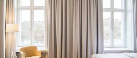 Hotellihuone, jossa on sänky, tuoli, lamppu ja suuret ikkunat, joista on näkymä ulos.