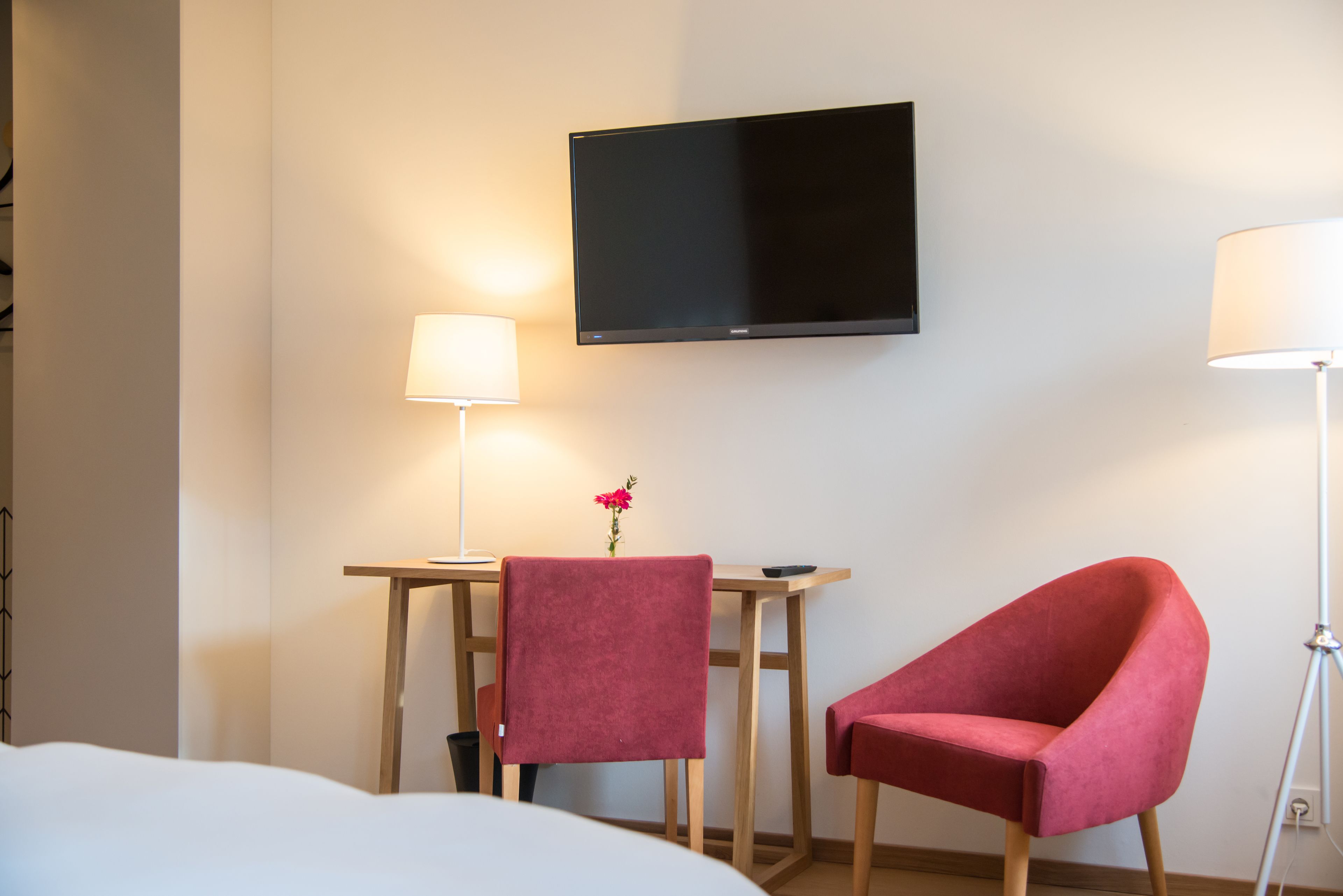 Una habitación de hotel con una cama, un escritorio con lámpara, un televisor y dos sillas rojas.