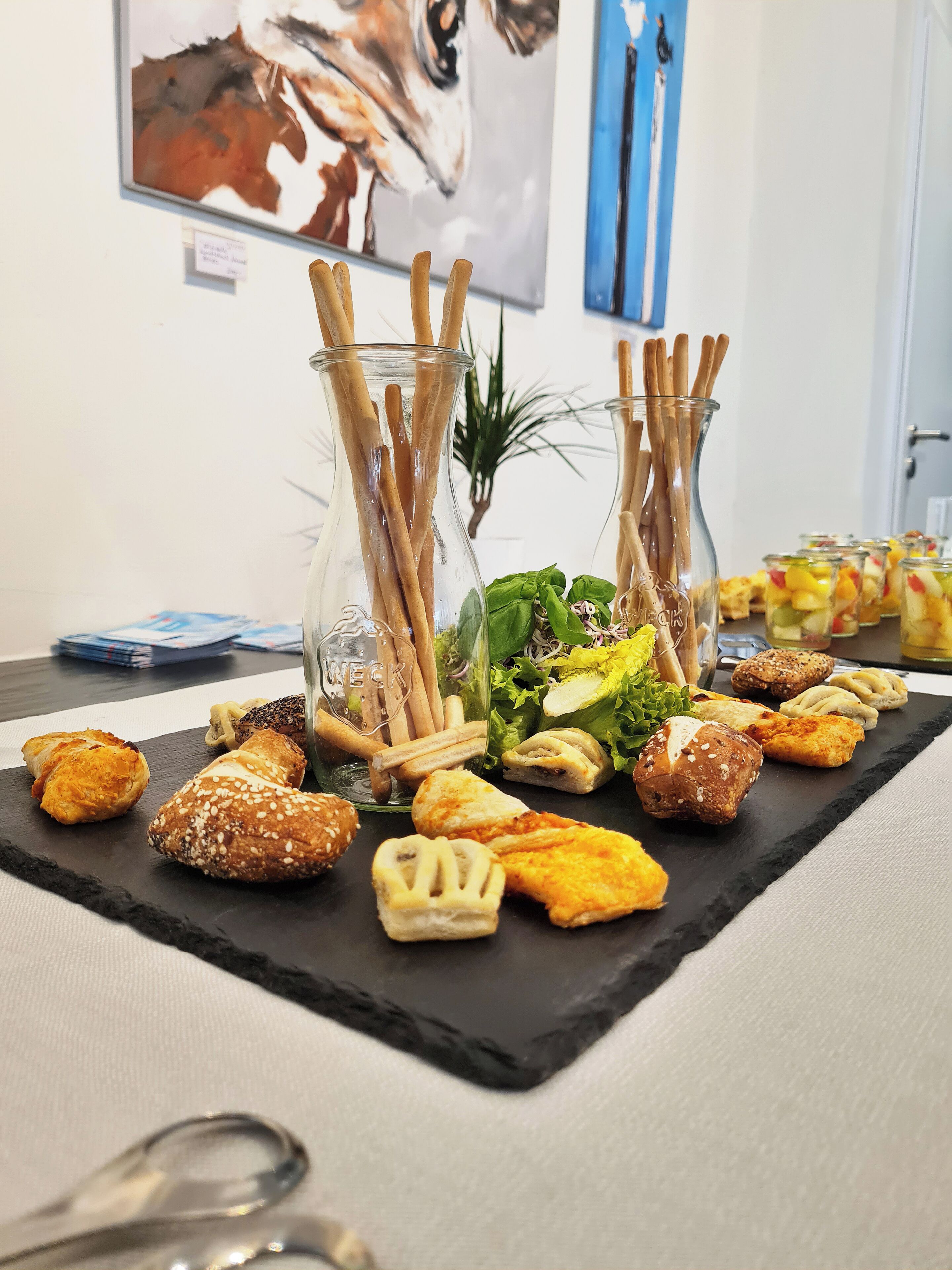 A table with various pastries and snacks, including croissants and sandwiches, in a room with artwork on the walls.