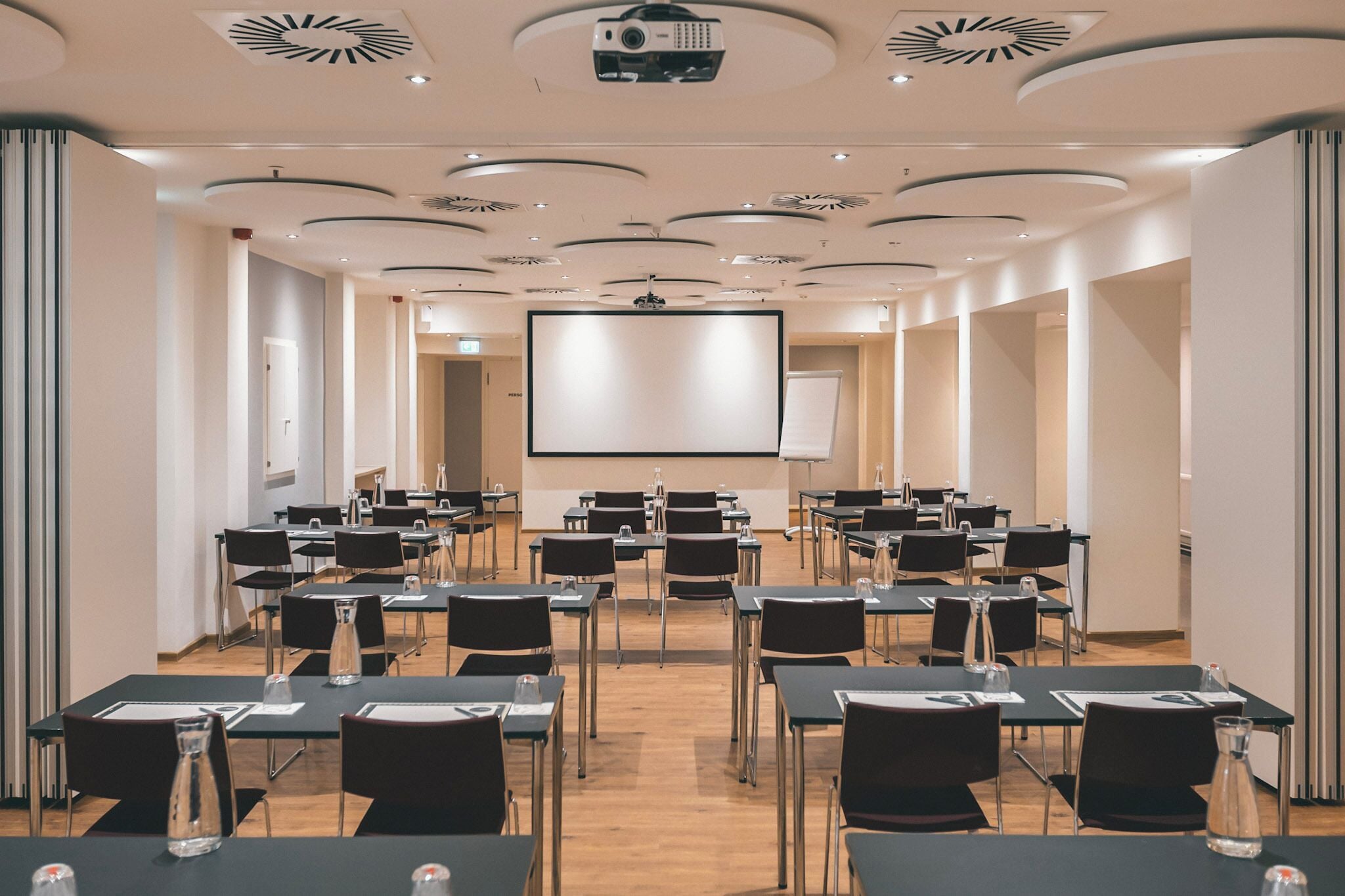 A conference room with rows of tables and chairs, a large screen, and a projector on the ceiling.