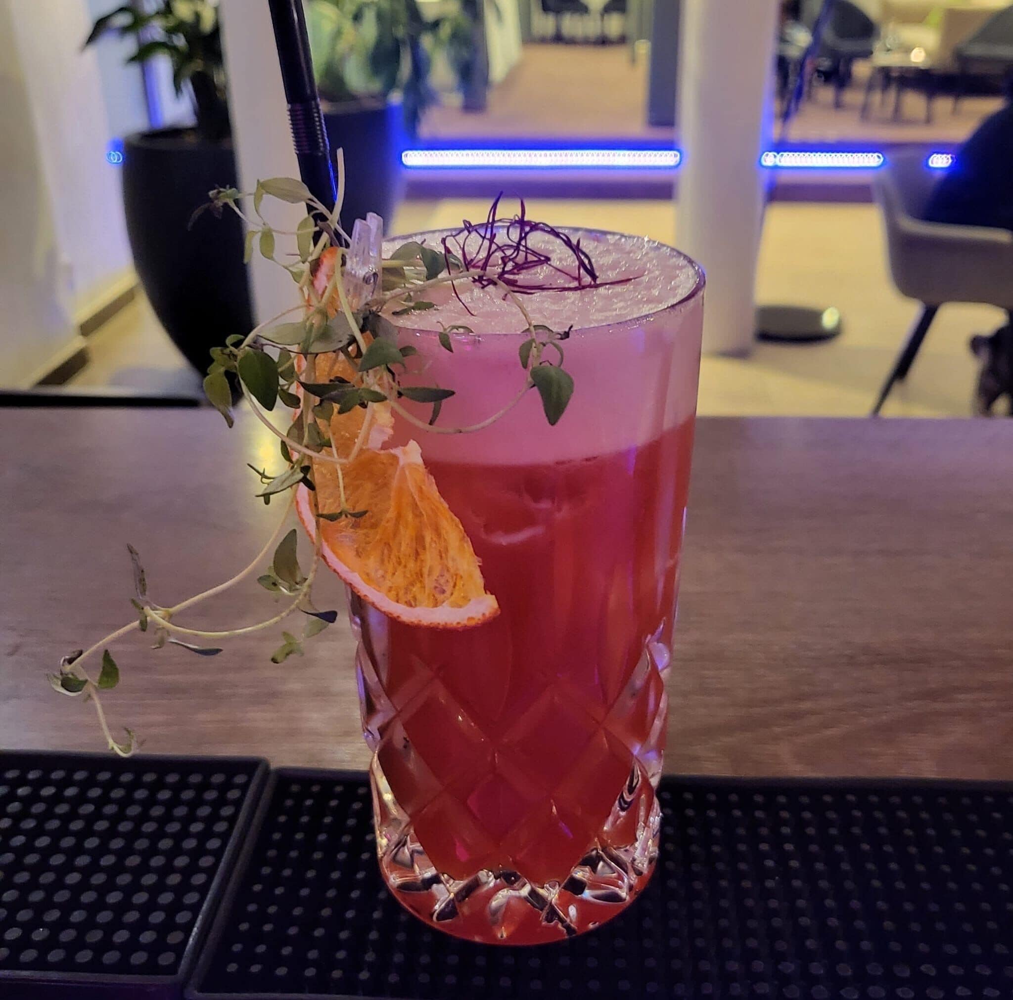 Een glas rode drank met een schijf sinaasappel en een takje groen op de rand, geplaatst op een baronderzetter.