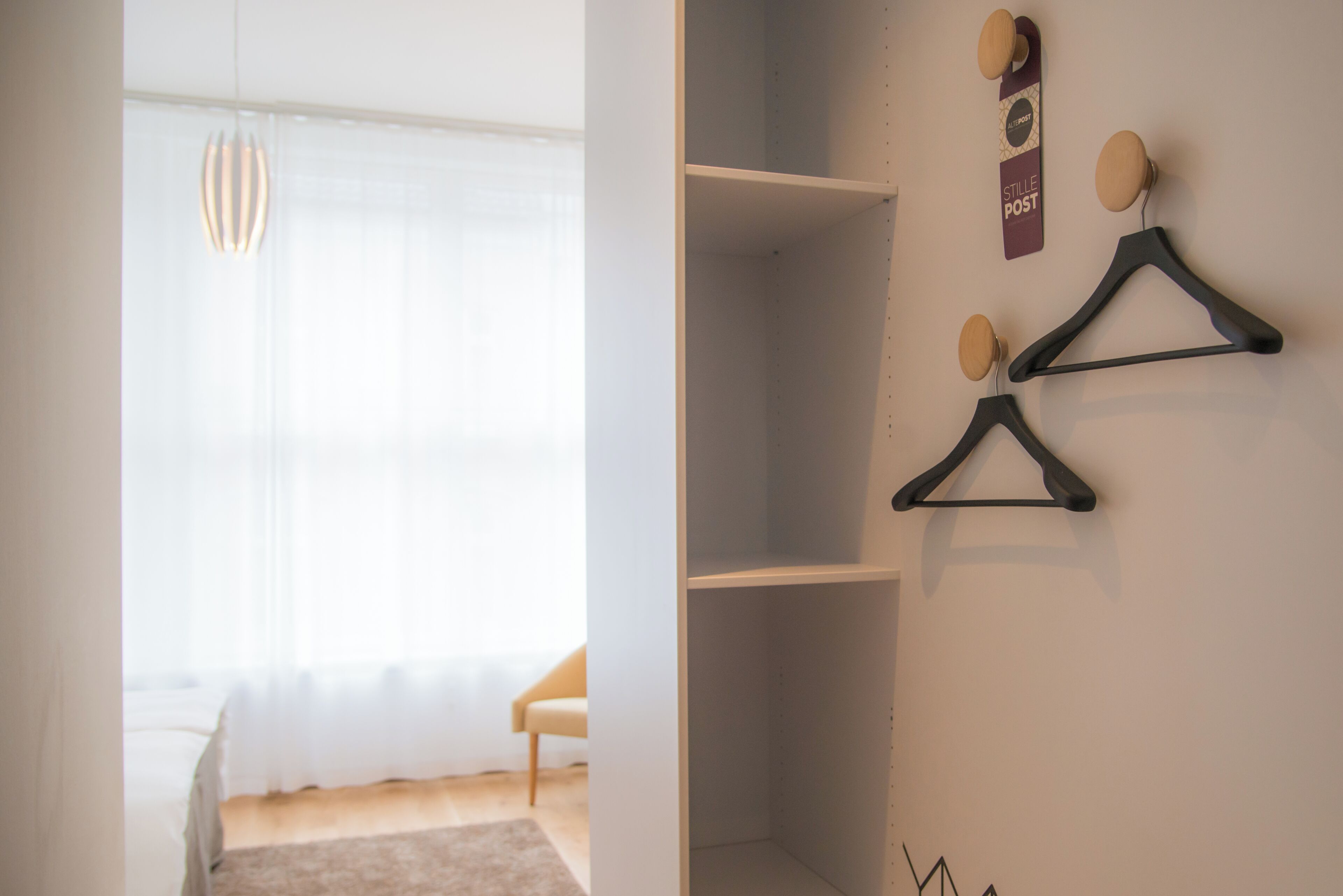 Une chambre avec un lit, une fenêtre ornée de rideaux légers, un luminaire suspendu et un placard contenant deux cintres noirs et une enseigne indiquant « STILE POST ».