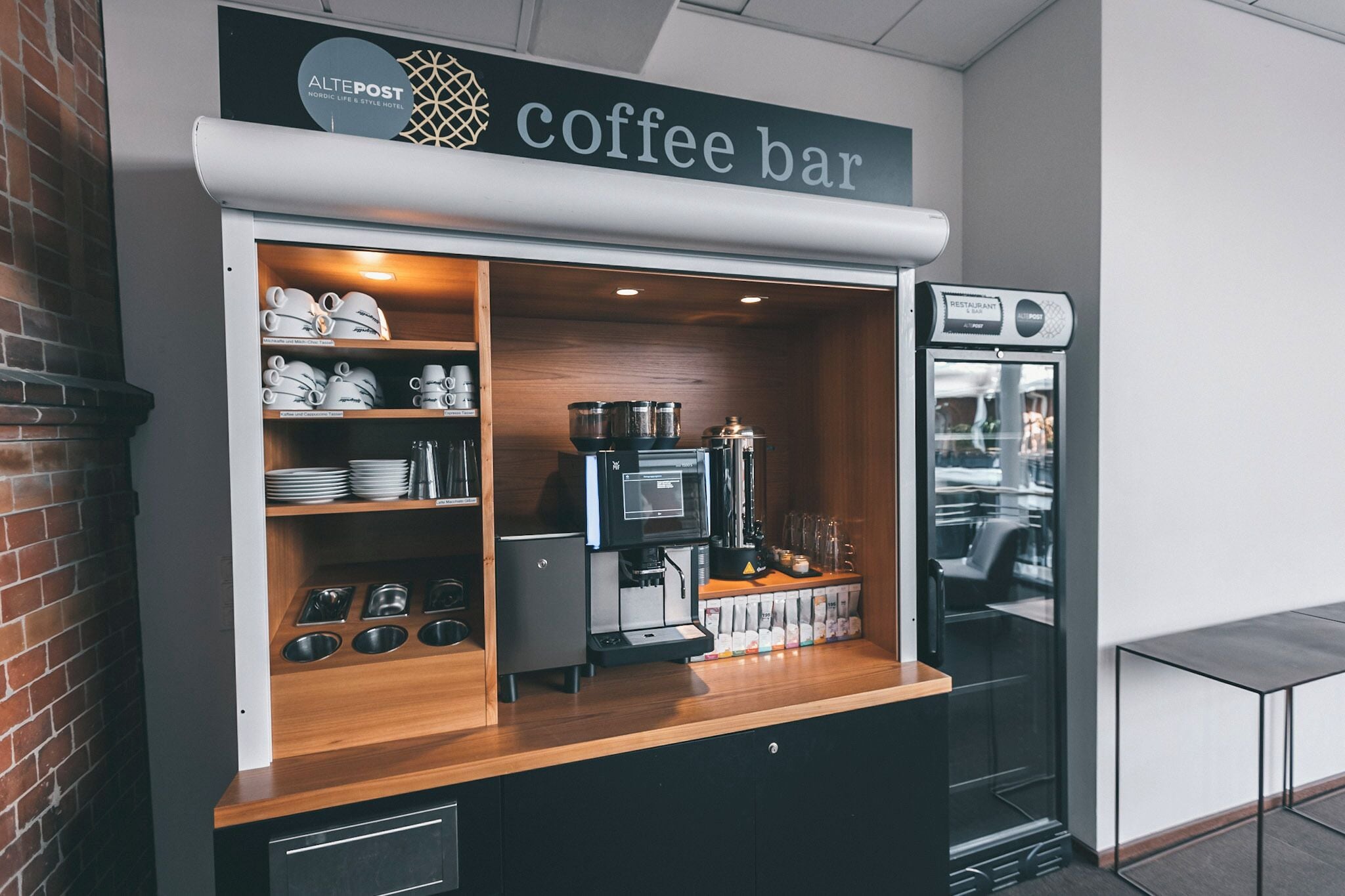 A coffee bar with a coffee machine, shelves with cups and saucers, and a refrigerator.