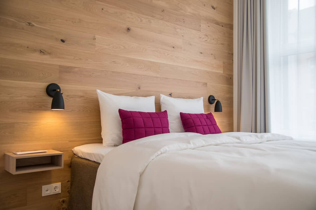 A modern bedroom with a wooden headboard, two bedside lamps, a bedside table, and a large bed with white bedding and two pink pillows.