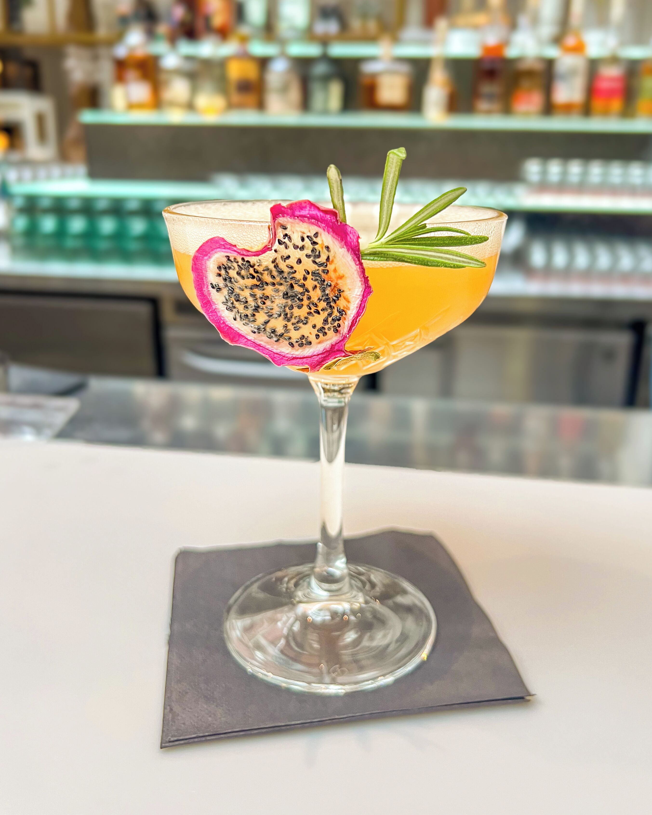 A cocktail glass with a heart-shaped slice of dragon fruit and a sprig of rosemary on a white table.