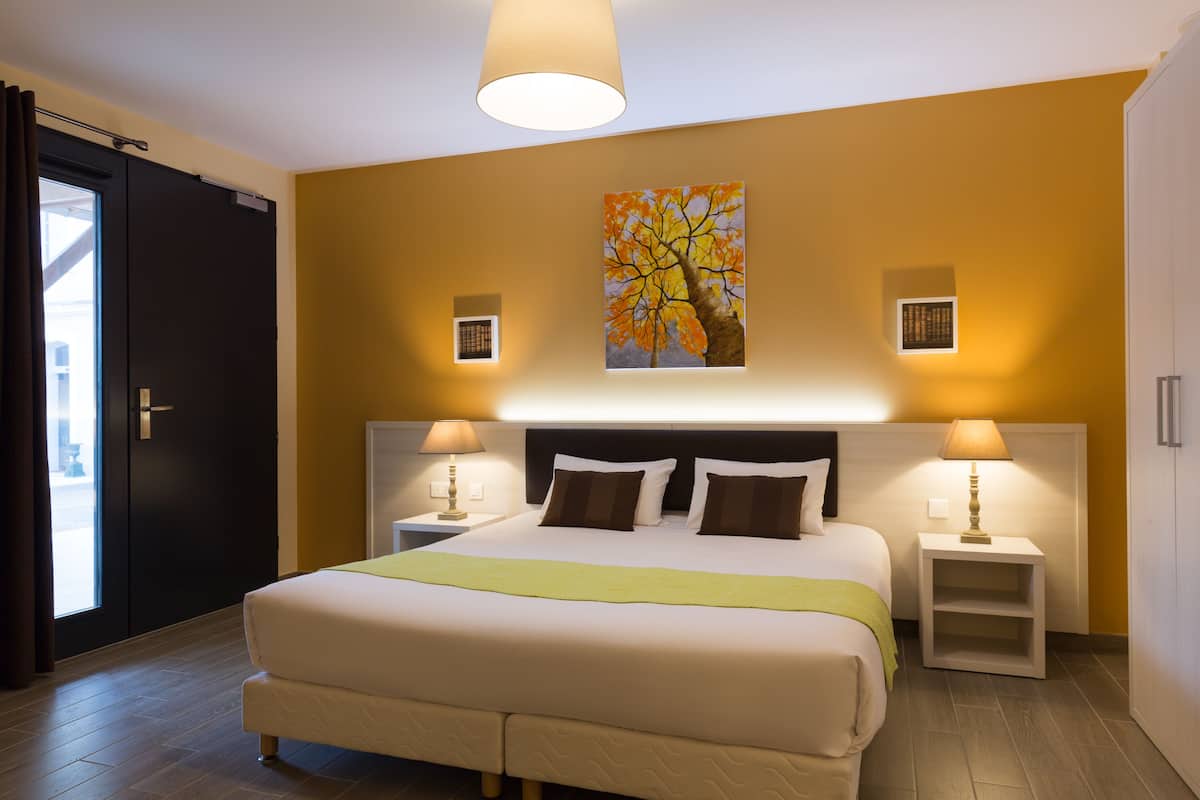 Una habitación de hotel con una cama grande, dos mesitas de noche con lámparas, un cuadro de un árbol y una puerta corredera.