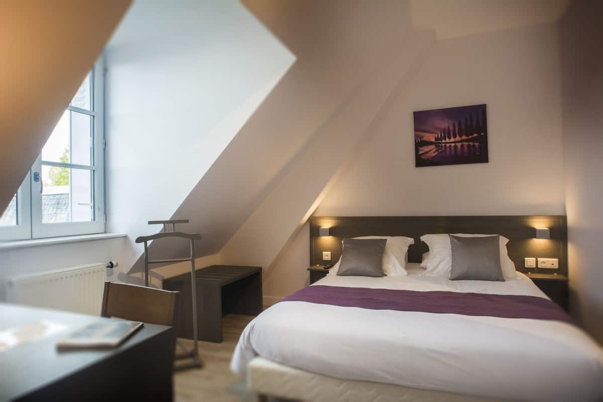 Una habitación de hotel moderna con techo inclinado, una cama grande con ropa blanca, un plaid morado y un cuadro de un paisaje en la pared.