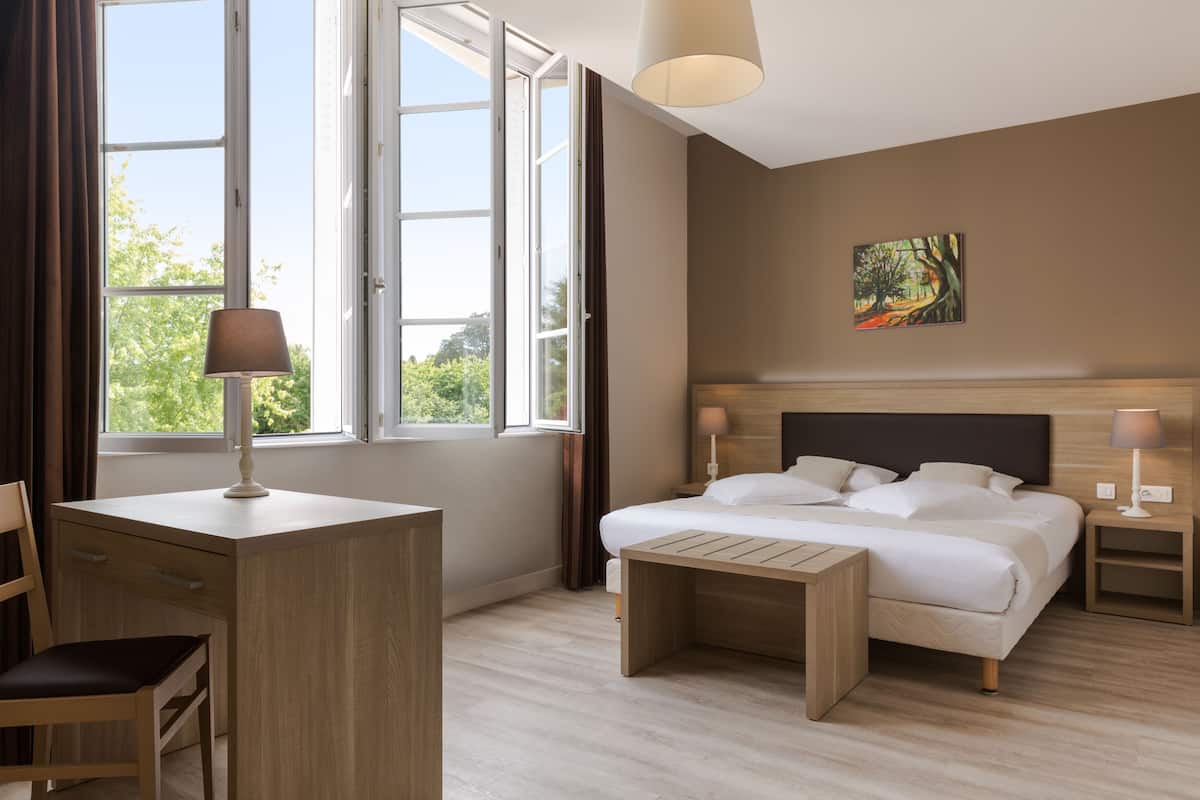 Un dormitorio con cama, mesitas de noche, un escritorio, una silla y una ventana con cortinas.