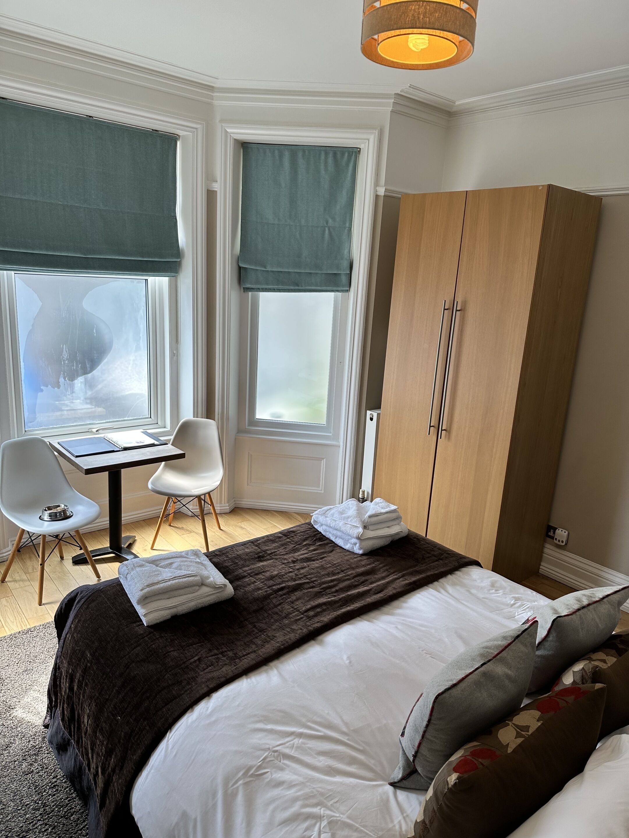 A bedroom with a bed, a desk with chairs, a wardrobe, and large windows with curtains.